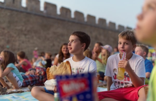 Kinder sitzen auf Decken im Freien in der Nähe einer Steinmauer, essen Snacks und trinken Getränke, während sie eine Veranstaltung oder Aufführung verfolgen.