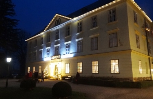 Ein großes, dreistöckiges, mit Lichterketten geschmücktes Gebäude in der Abenddämmerung. Mehrere Menschen sind in der Nähe eines Eingangs mit der Aufschrift "ADVENT" versammelt.