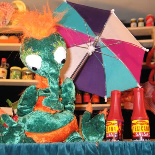 Ein grüner und orangefarbener Plüschdrache sitzt vor einem farbenfrohen Regenschirm und Salsaflaschen, während im Hintergrund Regale mit Lebensmitteln stehen.