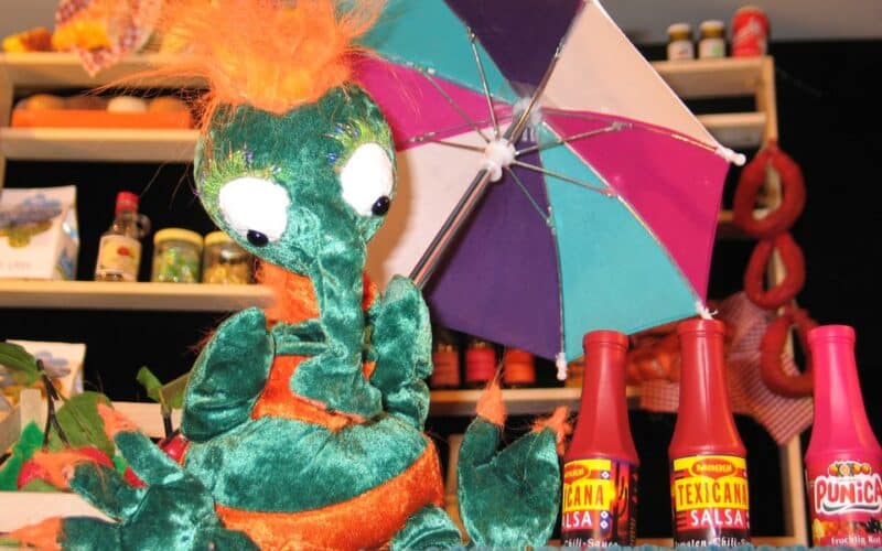 Ein grüner und orangefarbener Plüschdrache sitzt vor einem farbenfrohen Regenschirm und Salsaflaschen, während im Hintergrund Regale mit Lebensmitteln stehen.