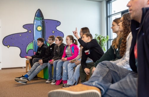 Eine Gruppe junger Leute sitzt in einer Reihe in einem Raum, einige halten Controller in der Hand, im Hintergrund ist eine Wandmalerei mit einer Cartoon-Rakete zu sehen.