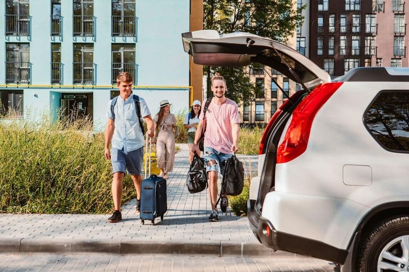Vier Personen mit Gepäck gehen auf einem gepflasterten Weg auf einen offenen Kofferraum zu, mit modernen Wohnhäusern und Grünflächen im Hintergrund.