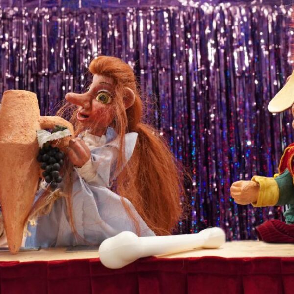 Drei farbenfrohe Puppen sitzen auf einer Bühne mit einem glänzenden lila Hintergrund, halten einen großen Krug und Weintrauben in der Hand und haben eine weiße Knochenstütze vor sich.