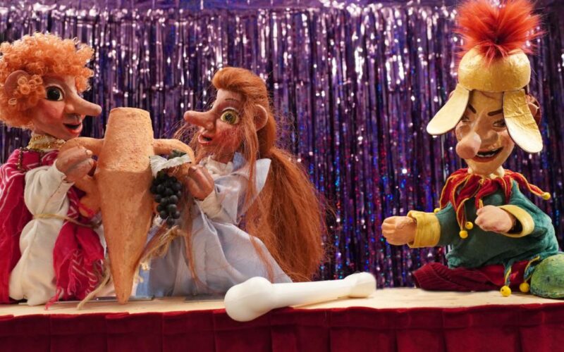 Drei farbenfrohe Puppen sitzen auf einer Bühne mit einem glänzenden lila Hintergrund, halten einen großen Krug und Weintrauben in der Hand und haben eine weiße Knochenstütze vor sich.