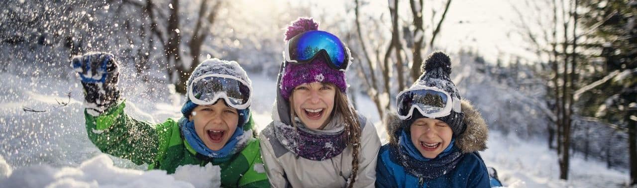 Drei Kinder in Winterkleidung und mit Skibrillen sitzen lächelnd und spielend im Schnee, mit Bäumen und Sonnenlicht im Hintergrund.