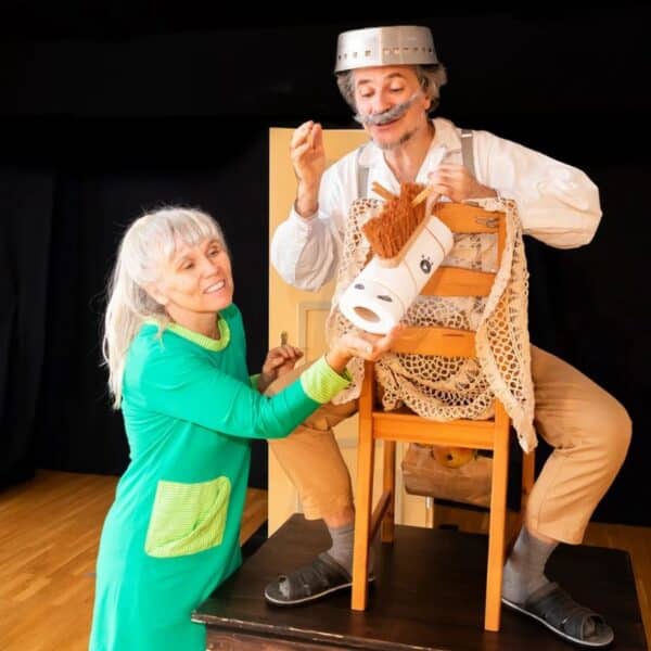 Zwei Erwachsene in Kostümen führen eine spielerische Szene auf; einer sitzt auf einem Stuhl und hat einen Topf auf dem Kopf, während der andere ihm eine Rolle Toilettenpapier reicht.