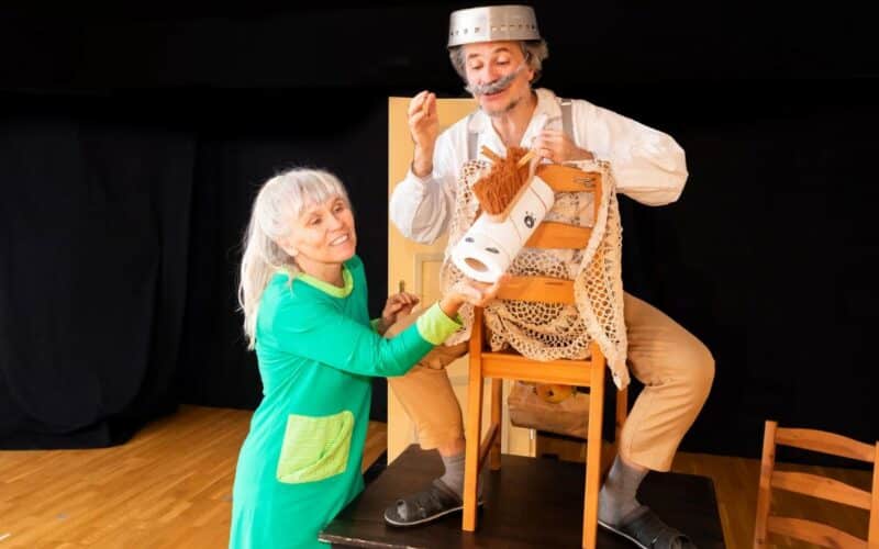 Zwei Erwachsene in Kostümen führen eine spielerische Szene auf; einer sitzt auf einem Stuhl und hat einen Topf auf dem Kopf, während der andere ihm eine Rolle Toilettenpapier reicht.