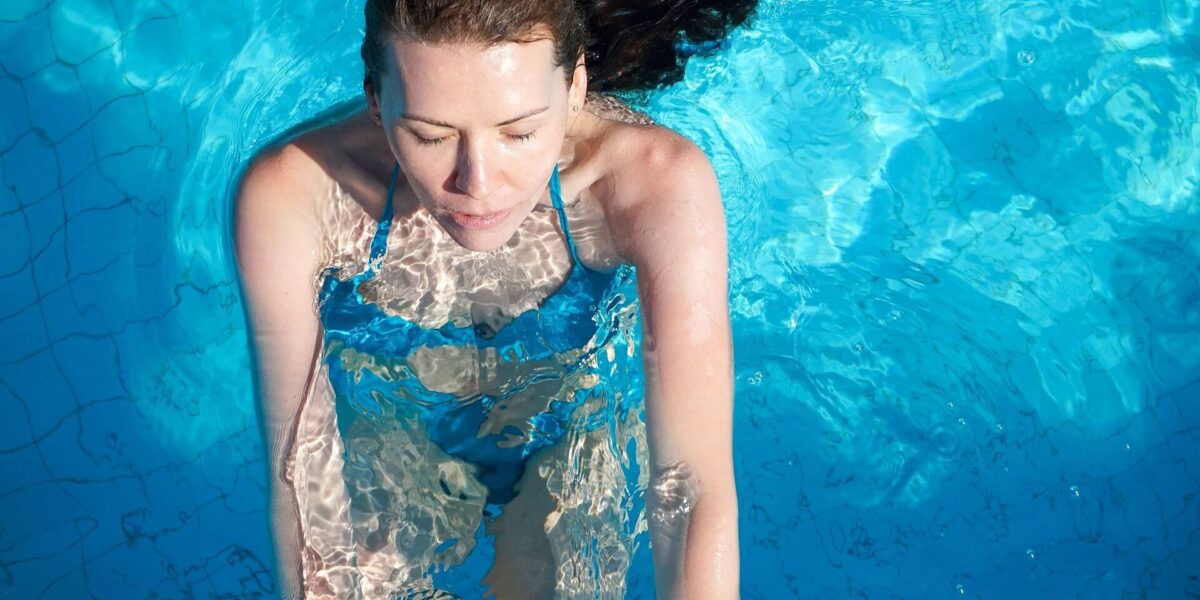 Eine Frau mit geschlossenen Augen steht aufrecht in einem Schwimmbecken mit blauem Wasser, ist teilweise untergetaucht und trägt einen blauen Badeanzug.