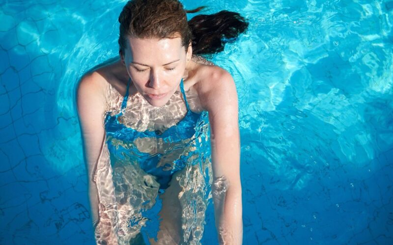 Eine Frau mit geschlossenen Augen steht aufrecht in einem Schwimmbecken mit blauem Wasser, ist teilweise untergetaucht und trägt einen blauen Badeanzug.