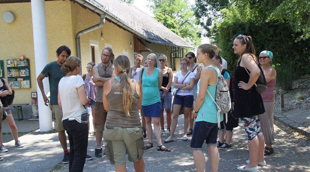 Eine Gruppe von Erwachsenen steht im Freien in einem Halbkreis in der Nähe eines Gebäudes und hört einer Person zu, die spricht. Einige Personen tragen Sonnenbrillen und Sommerkleidung. Bäume und eine Anzeigetafel sind zu sehen.