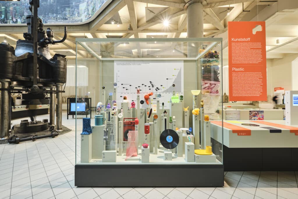 In einer Glasvitrine werden verschiedene farbenfrohe Kunststoffobjekte in einer modernen Museumsumgebung ausgestellt, rechts daneben befindet sich eine Informationstafel zum Thema Kunststoff.
