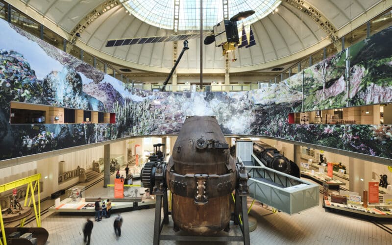 Eine große Museumsausstellung zeigt ein verrostetes U-Boot in der Mitte, umgeben von verschiedenen Maschinen und Ausstellungsstücken unter einer Glaskuppel und mit Wandmalereien bedeckten Wänden.