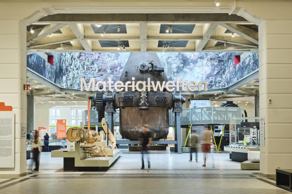Ausstellungsraum des Museums mit dem Wort "Materialwelten" über einer großen Industriemaschine; im Vordergrund sind Vitrinen und unscharfe Besucher zu sehen.
