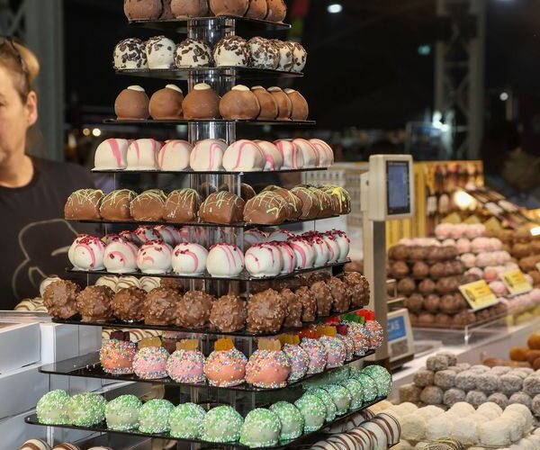 Ein mit verschiedenen Schokoladentrüffeln gefüllter Ständer an einem Marktstand, mit weiteren Pralinen und Lichtern im Hintergrund.