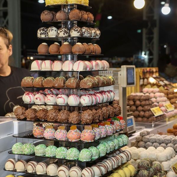 Ein mit verschiedenen Schokoladentrüffeln gefüllter Ständer an einem Marktstand, mit weiteren Pralinen und Lichtern im Hintergrund.