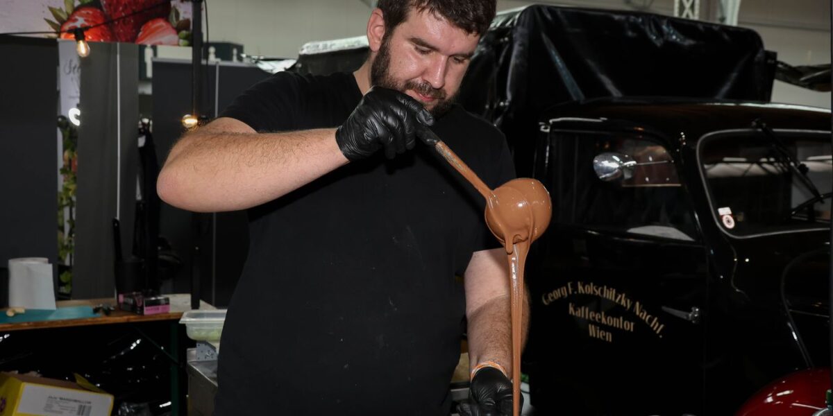 Ein Mann mit schwarzen Handschuhen gießt geschmolzene Schokolade aus einer Schöpfkelle auf einen Spatel bei einer Indoor-Veranstaltung mit verschiedenen Geräten und einem Oldtimer im Hintergrund.