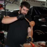 Ein Mann mit schwarzen Handschuhen gießt geschmolzene Schokolade aus einer Schöpfkelle auf einen Spatel bei einer Indoor-Veranstaltung mit verschiedenen Geräten und einem Oldtimer im Hintergrund.