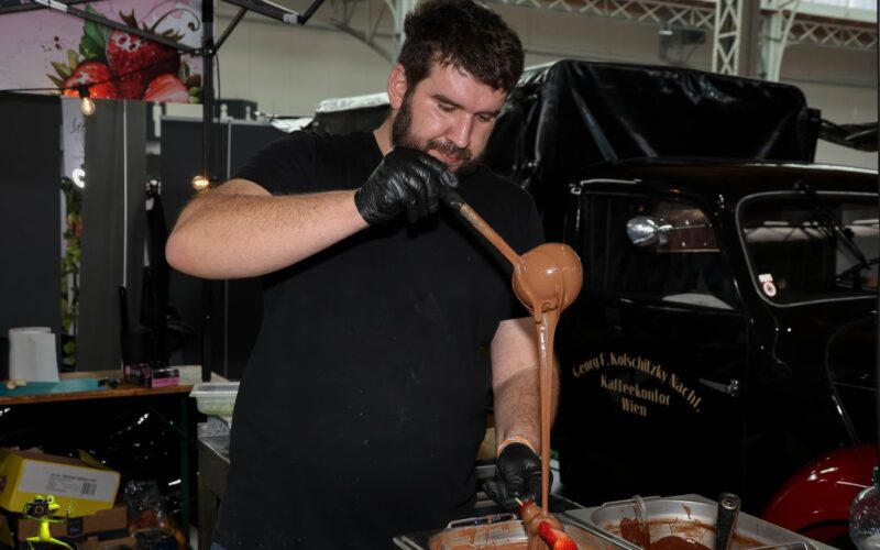 Ein Mann mit schwarzen Handschuhen gießt geschmolzene Schokolade aus einer Schöpfkelle auf einen Spatel bei einer Indoor-Veranstaltung mit verschiedenen Geräten und einem Oldtimer im Hintergrund.