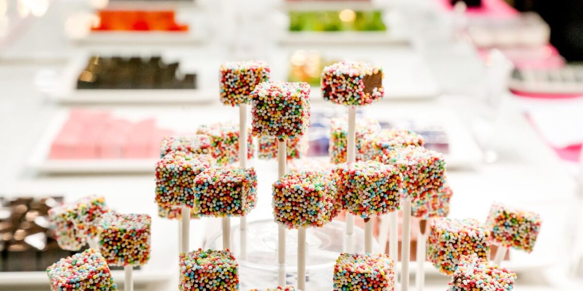 Bunte, mit Regenbogenstreuseln bedeckte Candy Pops werden auf Stäben präsentiert, während im Hintergrund Tabletts mit verschiedenen Süßigkeiten verschwimmen.