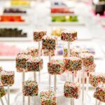 Bunte, mit Regenbogenstreuseln bedeckte Candy Pops werden auf Stäben präsentiert, während im Hintergrund Tabletts mit verschiedenen Süßigkeiten verschwimmen.