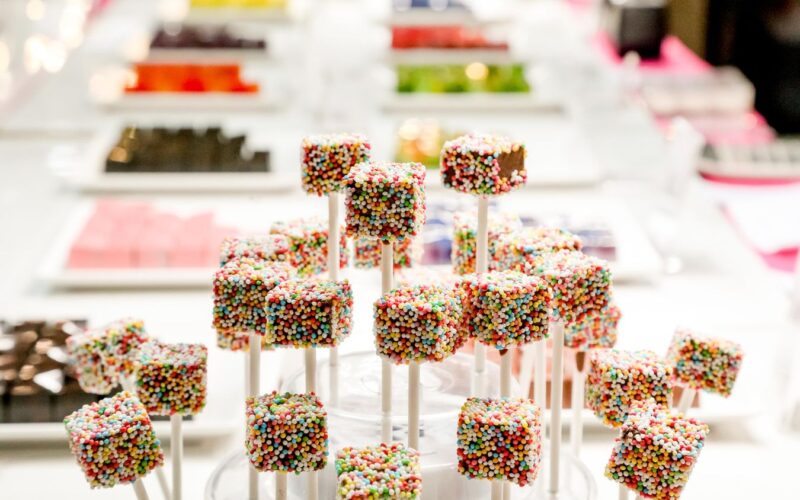 Bunte, mit Regenbogenstreuseln bedeckte Candy Pops werden auf Stäben präsentiert, während im Hintergrund Tabletts mit verschiedenen Süßigkeiten verschwimmen.