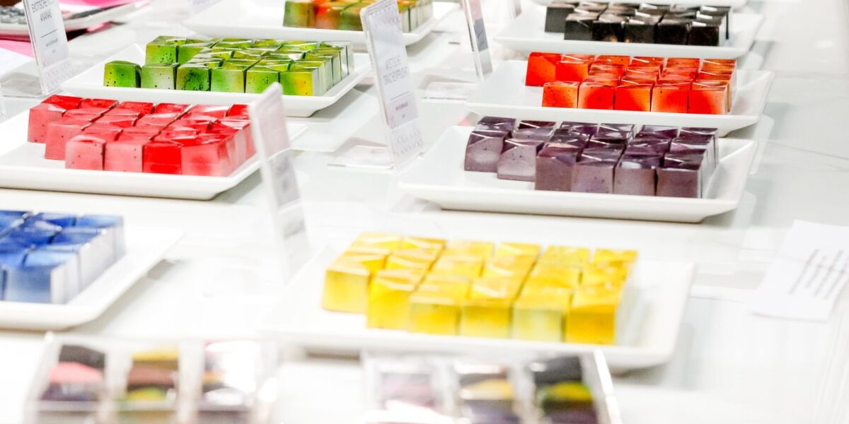Auf einer weißen Theke stehen Teller mit bunten, ordentlich angeordneten, quadratischen Desserts.