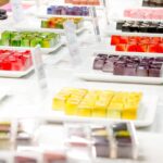 Auf einer weißen Theke stehen Teller mit bunten, ordentlich angeordneten, quadratischen Desserts.