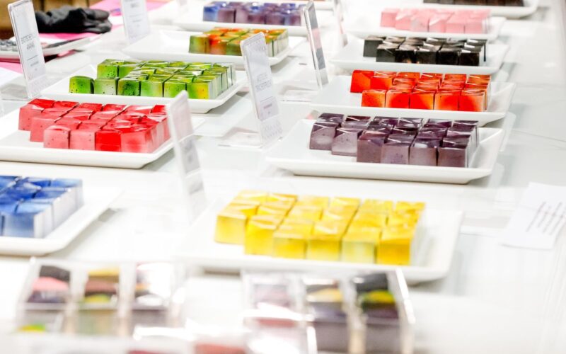 Auf einer weißen Theke stehen Teller mit bunten, ordentlich angeordneten, quadratischen Desserts.