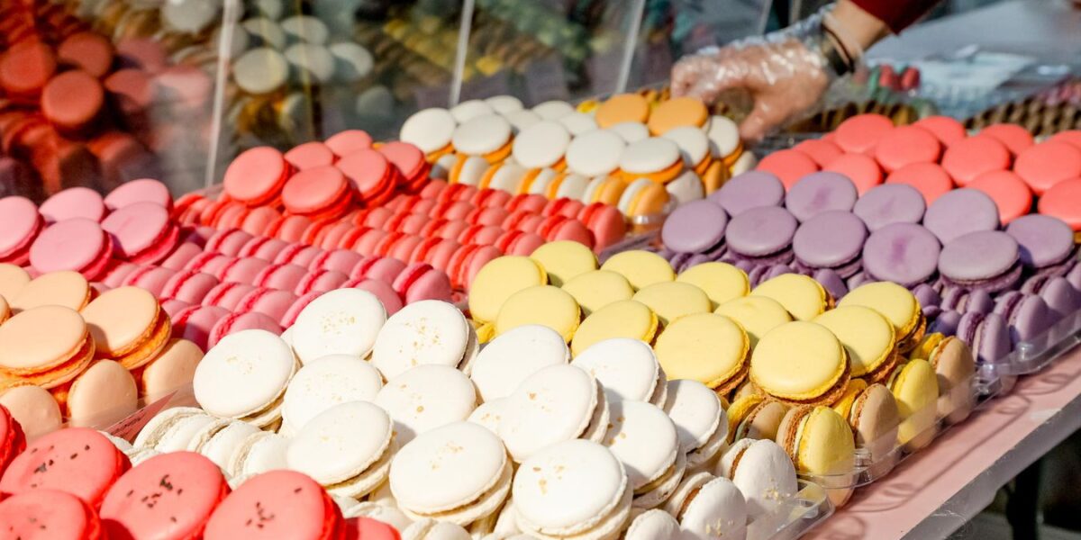 Eine Vielzahl von bunten Macarons ist in Reihen auf einer Theke ausgestellt, und eine behandschuhte Hand greift hinter einer durchsichtigen Trennwand nach ihnen.