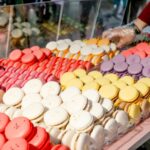 Eine Vielzahl von bunten Macarons ist in Reihen auf einer Theke ausgestellt, und eine behandschuhte Hand greift hinter einer durchsichtigen Trennwand nach ihnen.