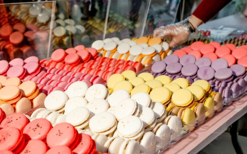 Eine Vielzahl von bunten Macarons ist in Reihen auf einer Theke ausgestellt, und eine behandschuhte Hand greift hinter einer durchsichtigen Trennwand nach ihnen.