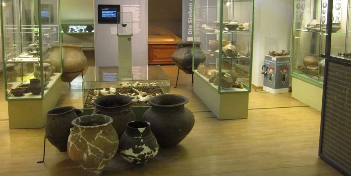 In Vitrinen und Regalen einer Museumsausstellung sind antike Töpferwaren, große Gefäße und Artefakte ausgestellt; im Hintergrund sind Knochen und Informationstafeln zu sehen.