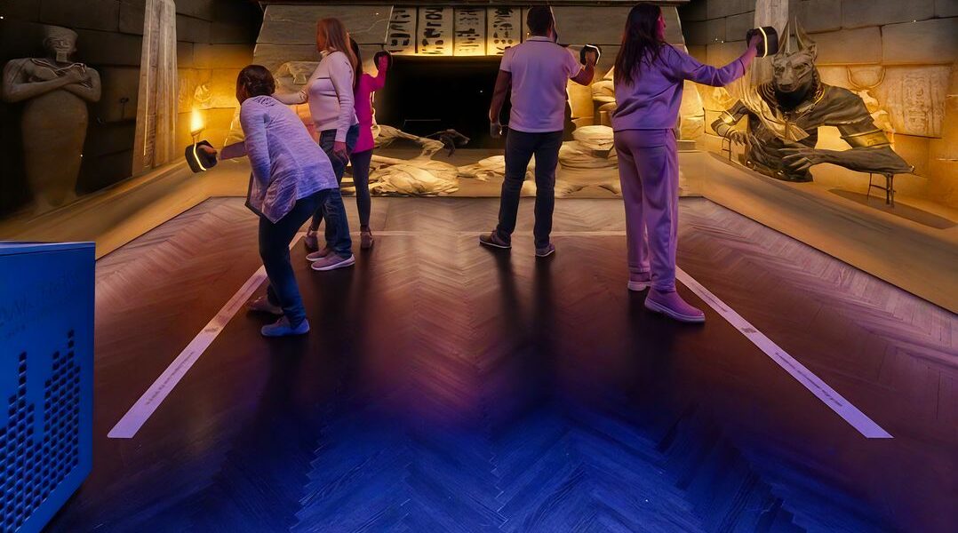 Fünf Personen mit Virtual-Reality-Headsets in einem Raum mit altägyptischem Dekor, darunter Statuen, Sarkophage und Hieroglyphen an den Wänden.