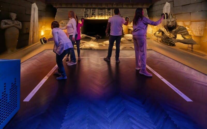 Fünf Personen mit Virtual-Reality-Headsets in einem Raum mit altägyptischem Dekor, darunter Statuen, Sarkophage und Hieroglyphen an den Wänden.