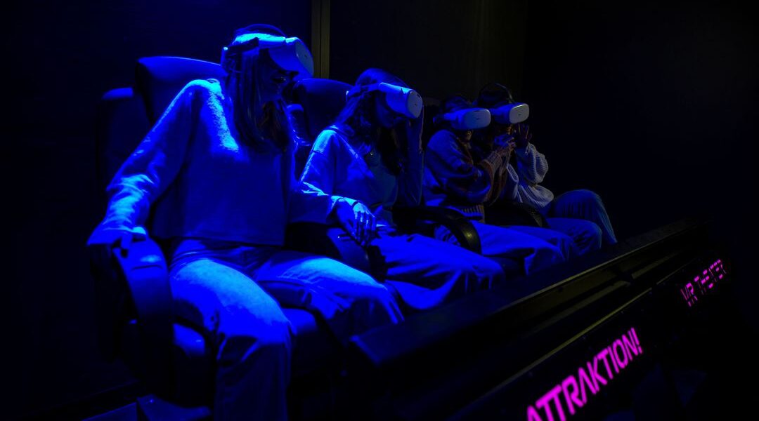 Vier Personen sitzen auf Stühlen und tragen Virtual-Reality-Headsets, die von blauem Licht in einem dunklen Raum beleuchtet werden. Das Wort "ATTRACTION" ist auf einem Schild in der Nähe zu lesen.
