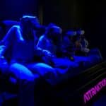Vier Personen sitzen auf Stühlen und tragen Virtual-Reality-Headsets, die von blauem Licht in einem dunklen Raum beleuchtet werden. Das Wort "ATTRACTION" ist auf einem Schild in der Nähe zu lesen.