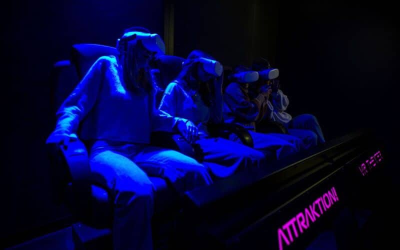 Vier Personen sitzen auf Stühlen und tragen Virtual-Reality-Headsets, die von blauem Licht in einem dunklen Raum beleuchtet werden. Das Wort "ATTRACTION" ist auf einem Schild in der Nähe zu lesen.