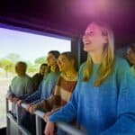 Eine Gruppe von Menschen steht an einem Geländer und schaut auf einen großen Bildschirm, der eine Naturszene mit Bäumen und Gras in einem Raum mit künstlicher Beleuchtung zeigt.