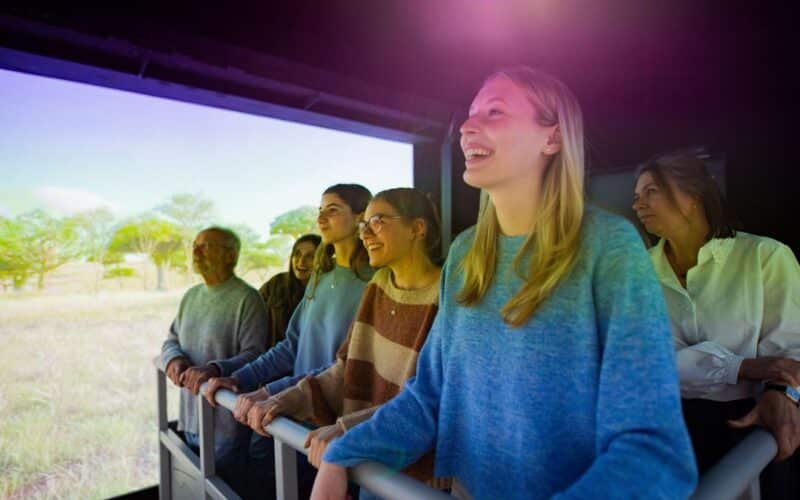 Eine Gruppe von Menschen steht an einem Geländer und schaut auf einen großen Bildschirm, der eine Naturszene mit Bäumen und Gras in einem Raum mit künstlicher Beleuchtung zeigt.