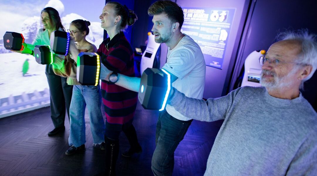 Vier Personen stehen in einer Reihe und halten leuchtende Controller in der Hand, die in einem schwach beleuchteten Raum mit einem interaktiven digitalen Display interagieren.