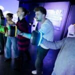 Vier Personen stehen in einer Reihe und halten leuchtende Controller in der Hand, die in einem schwach beleuchteten Raum mit einem interaktiven digitalen Display interagieren.