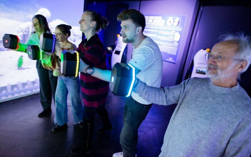 Vier Personen stehen in einer Reihe und halten leuchtende Controller in der Hand, die in einem schwach beleuchteten Raum mit einem interaktiven digitalen Display interagieren.