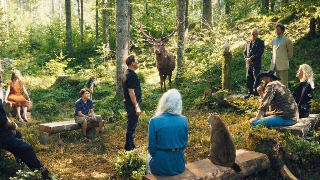 Eine Gruppe von Menschen und zwei Tieren sitzt auf Bänken in einer Waldlichtung, gegenüber einem stehenden Mann und einem großen Hirsch in der Mitte.