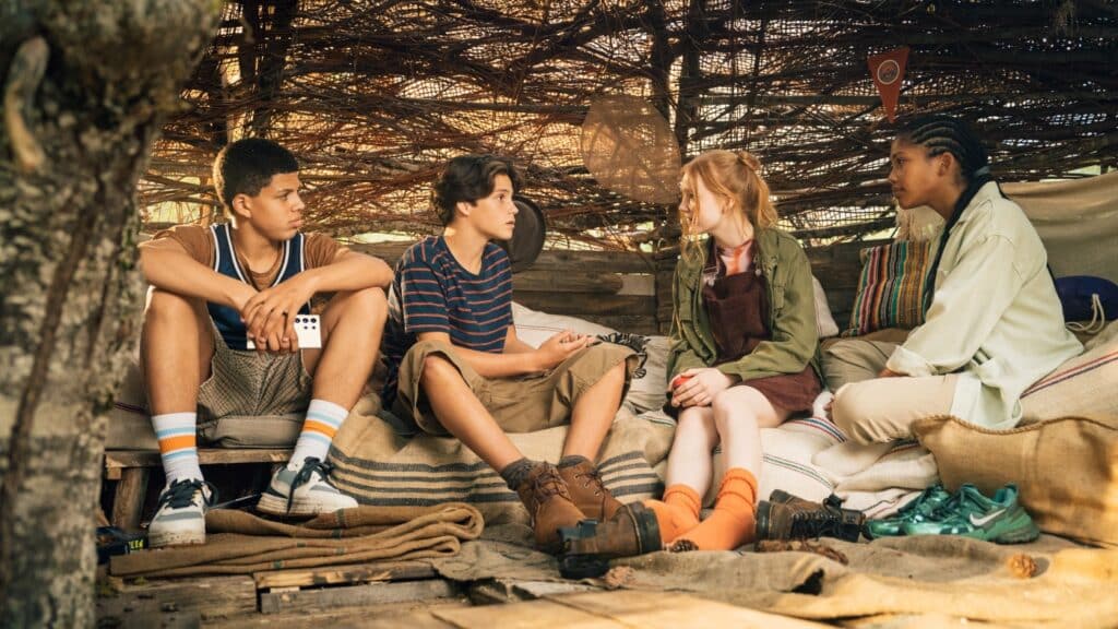 Vier Kinder sitzen in einem rustikalen, behelfsmäßigen Unterstand aus Ästen und unterhalten sich, umgeben von Decken und Kissen.