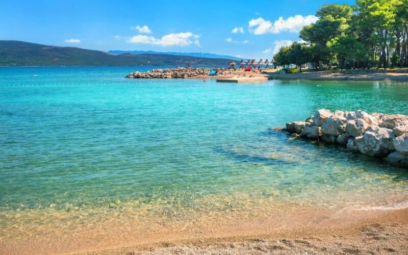 Klares türkisfarbenes Wasser trifft auf einen Sandstrand mit Felsen im Vordergrund; Sonnenanbeter entspannen sich auf Stühlen unter Sonnenschirmen in der Nähe von Bäumen im Hintergrund unter einem blauen Himmel.