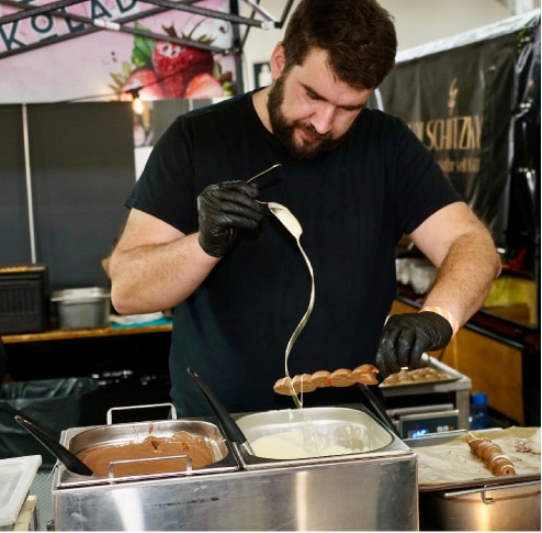 Ein Mann mit schwarzen Handschuhen träufelt weiße Schokolade über aufgespießte Leckereien an einem Marktstand mit Schokoladenbehältern vor ihm.
