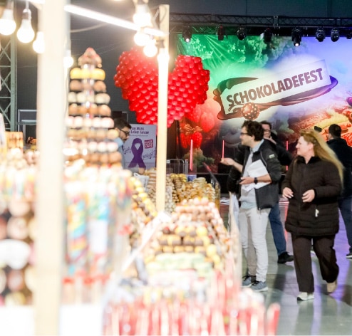 Menschen gehen an Ständen mit verschiedenen Süßigkeiten vorbei, die auf einer Indoor-Veranstaltung mit einem "Schokoladefest"-Schild und einem großen roten Herz aus Luftballons im Hintergrund angeboten werden.