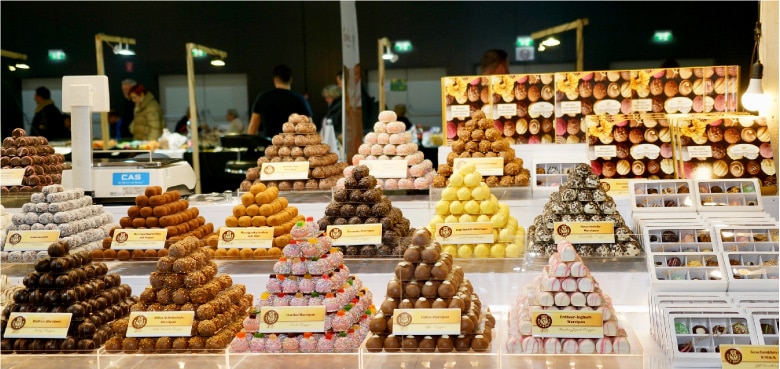 Auslage verschiedener pyramidenförmiger Tabletts, gefüllt mit verschiedenen bunten Schokoladentrüffeln und Süßigkeiten an einem Marktstand, mit Menschen und Lichtern im Hintergrund.