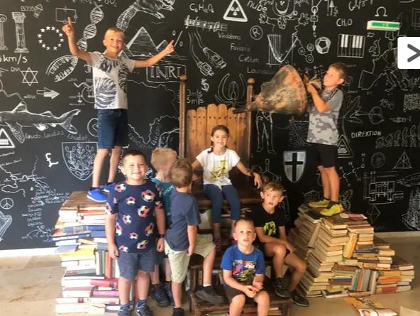 Eine Gruppe von Kindern posiert auf und um einen Holzstuhl, der auf einem Bücherstapel steht, während im Hintergrund eine Kreidetafel mit naturwissenschaftlichen und mathematischen Zeichnungen und Formeln zu sehen ist.