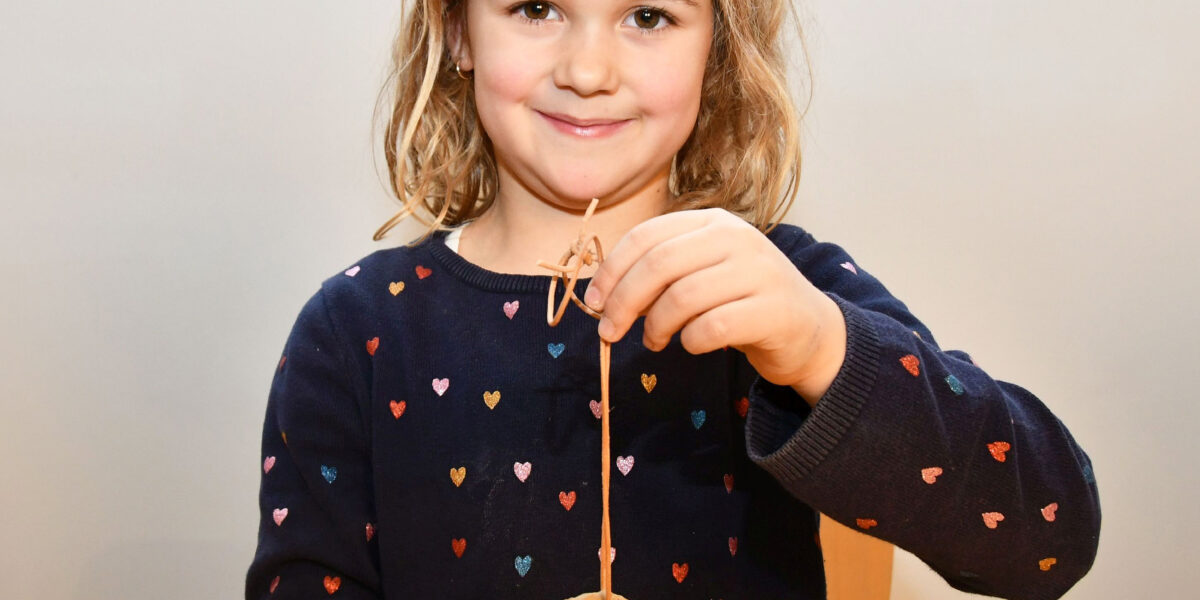 Ein junges Mädchen mit hellbraunem Haar hält ein rundes, verziertes Handwerk aus Ton an einer Schnur hoch. Sie trägt einen marineblauen Pullover mit bunten Herzmustern.